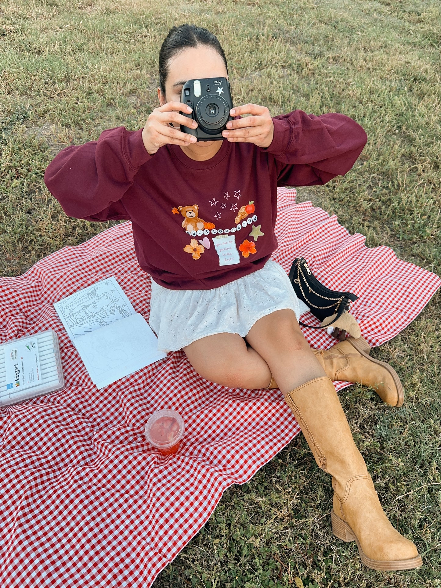 Person sitting on a red checkered blanket with a maroon scrapbook inspired sweatshirt and skirt, holding a camera to give an aesthetic look!